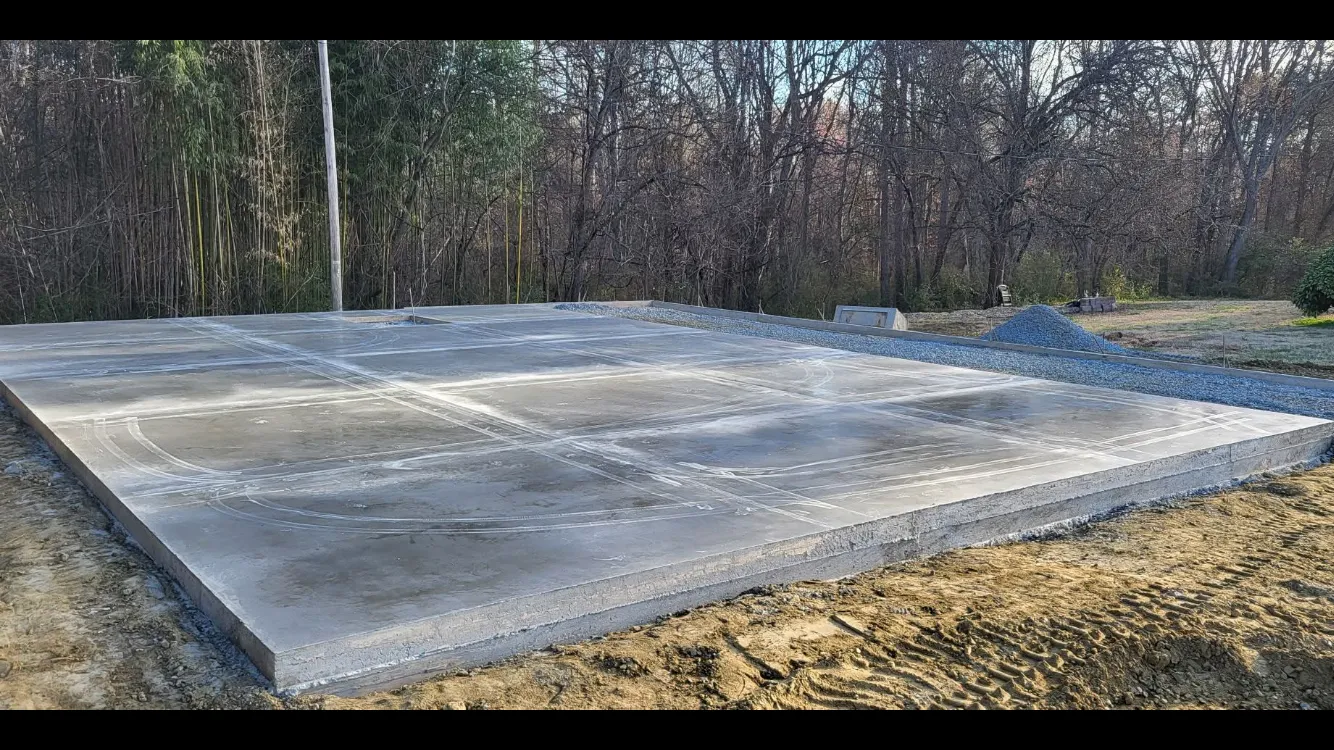 Concrete foundation slab, rectangular with divided sections, surrounded by dirt and gravel; trees in the background.