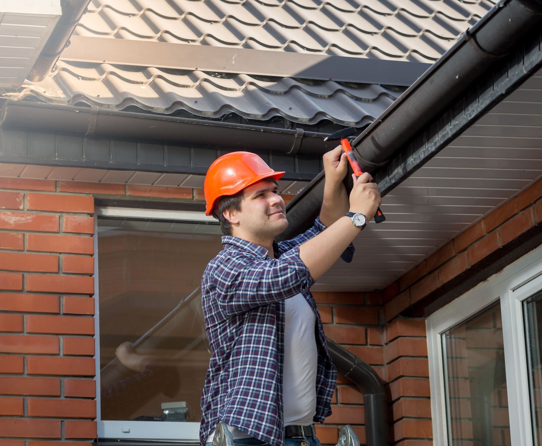 Worker in orange hard hat tightening gutter on a brick house roof edge