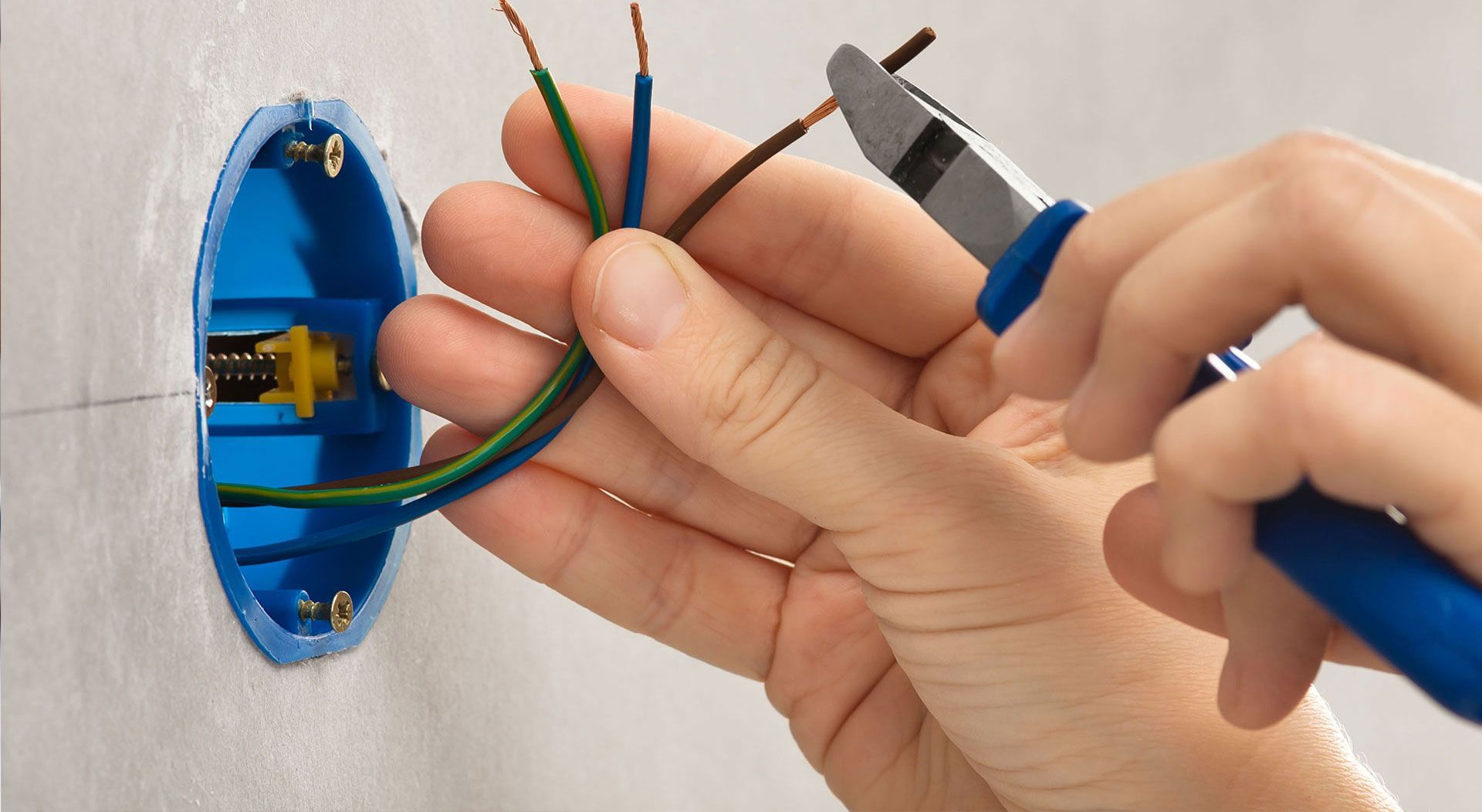 Hands stripping electrical wires in a blue wall junction box with pliers