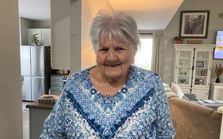 An elderly woman is sitting in a living room wearing a blue and white shirt.