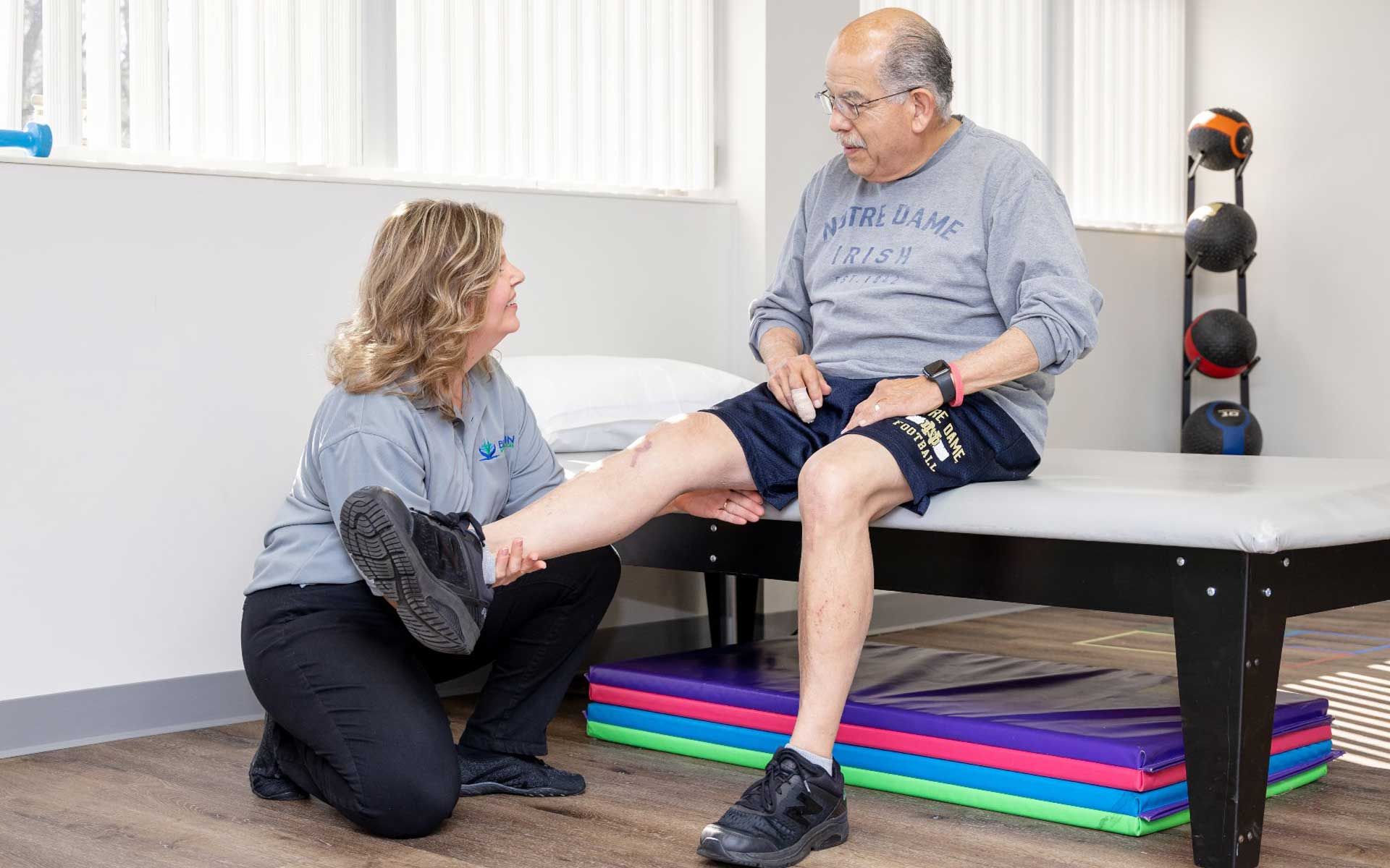 A woman is kneeling down to help an older man stretch his leg.