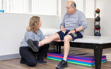 A woman is kneeling down to help an older man stretch his leg.