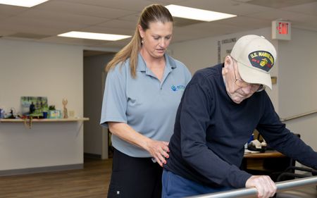 A woman is helping an older man walk on a parallel bars