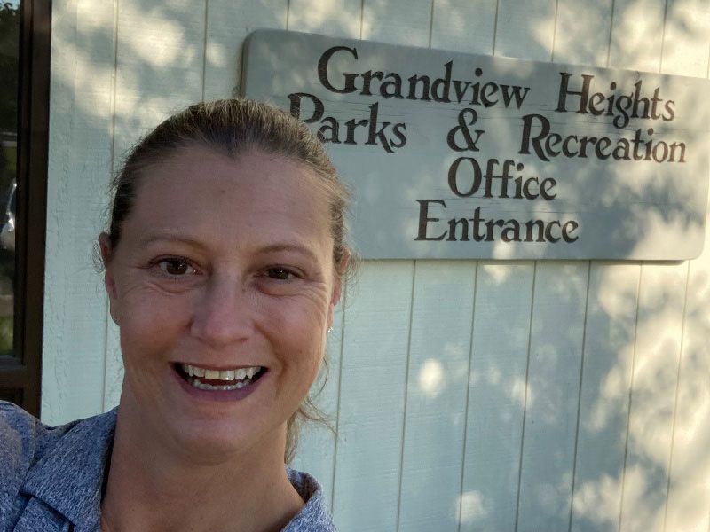 A woman is smiling in front of a sign for grandview heights parks and recreation office entrance