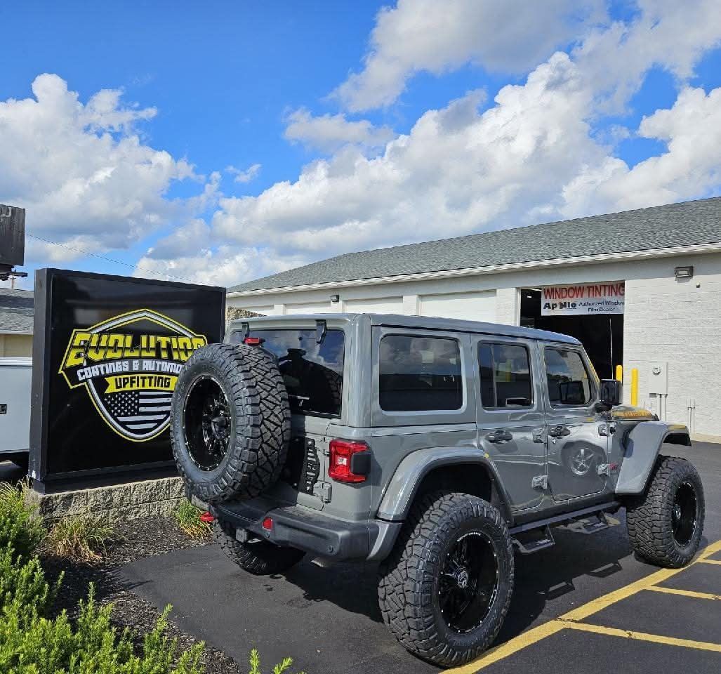 Gray Jeep with black wheels parked in front of a sign for Evolution Customs and a building under a blue sky.