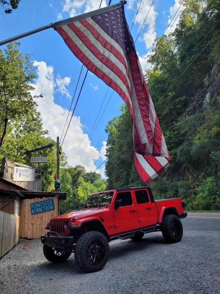 Red Jeep under American flag near a roadside building and trees, on a sunny day.