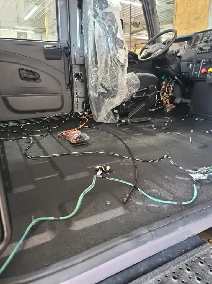 Interior of a truck cab under construction; exposed wiring, seat covered in plastic, gray floor.