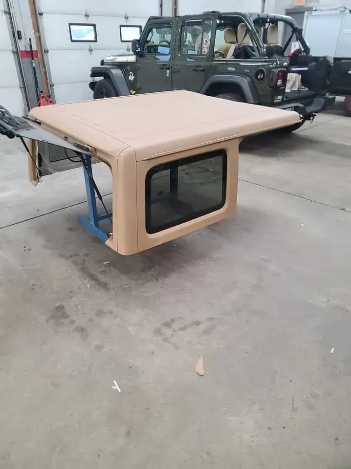 Tan Jeep hardtop on a stand with a green Jeep in the background.