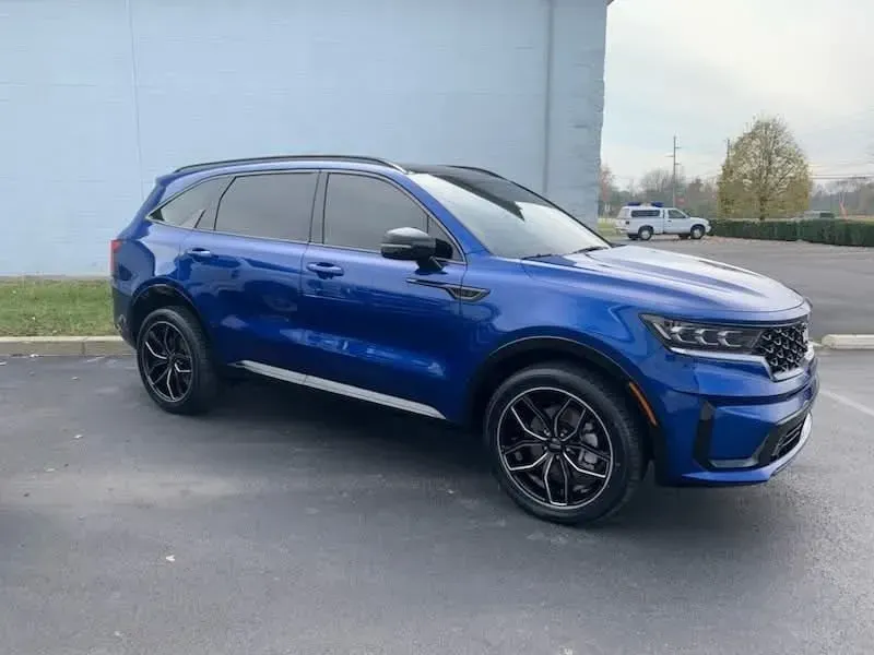 Blue Kia Sorento SUV parked on asphalt. Black wheels, tinted windows, and a light blue wall.