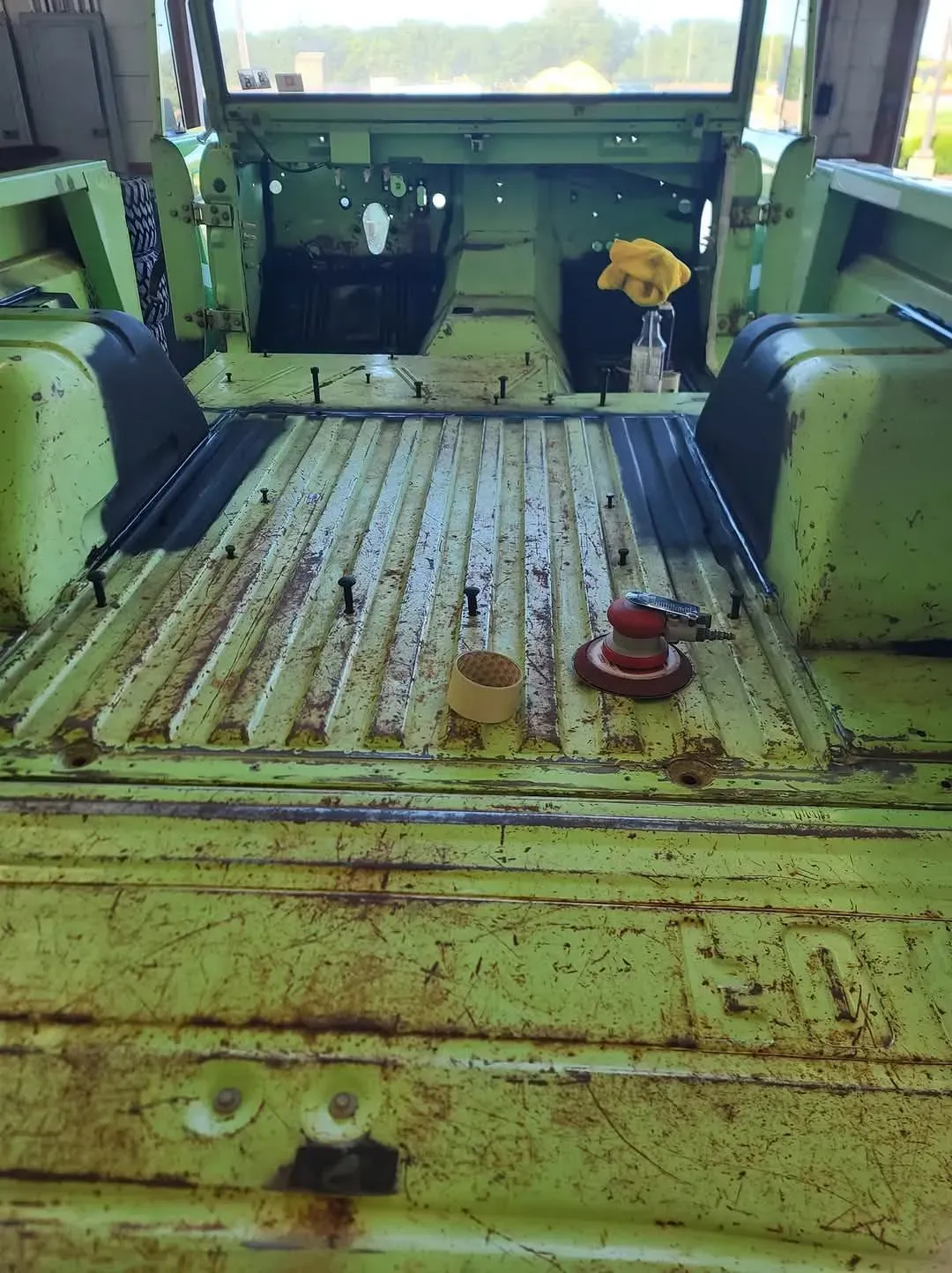 Interior of a light green vintage vehicle. Rusted bed with tools. Yellow flowers in a jar.