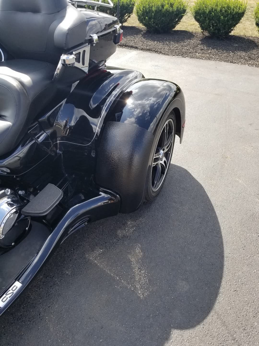 Black trike motorcycle parked on asphalt. Rear wheel and fender visible, with shiny surfaces and a shadow.
