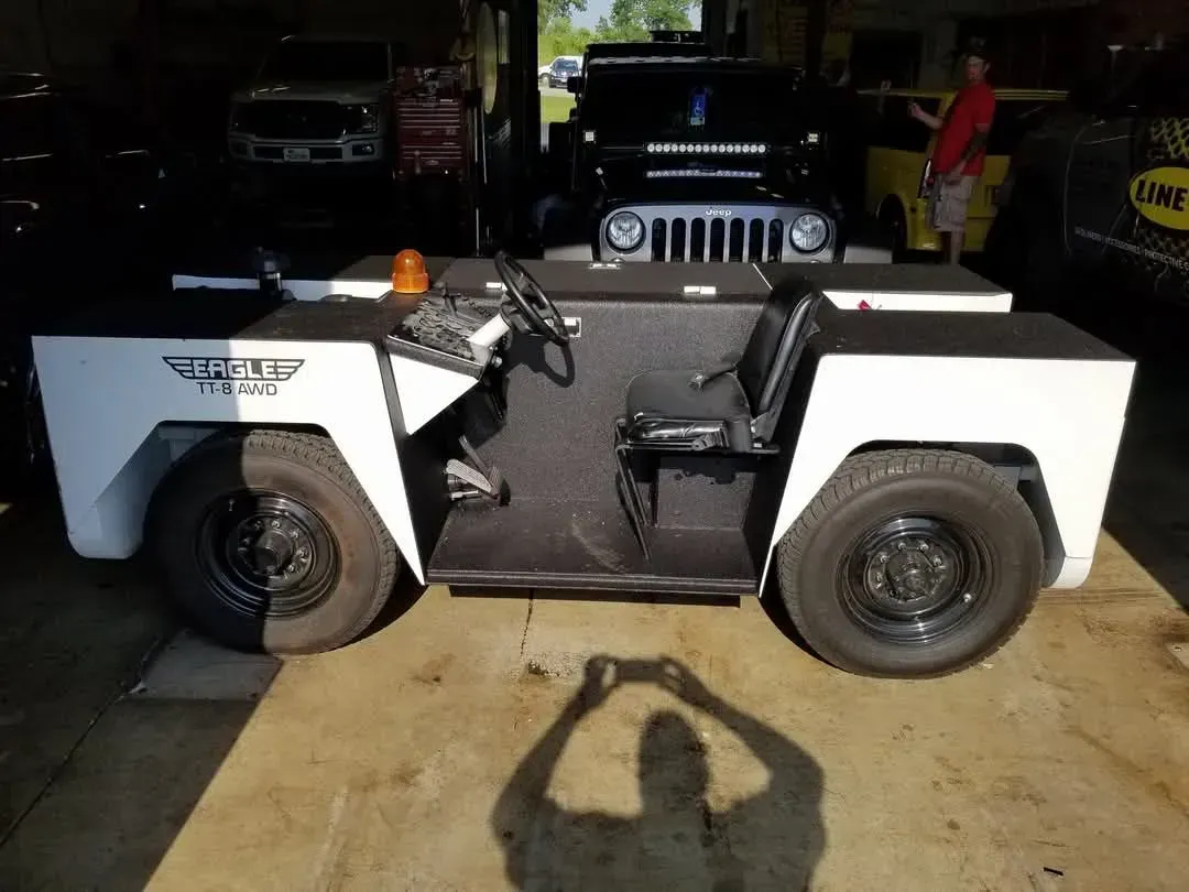White industrial tug vehicle inside a garage. Driver's seat, dashboard, steering wheel visible. Person taking photo reflected.
