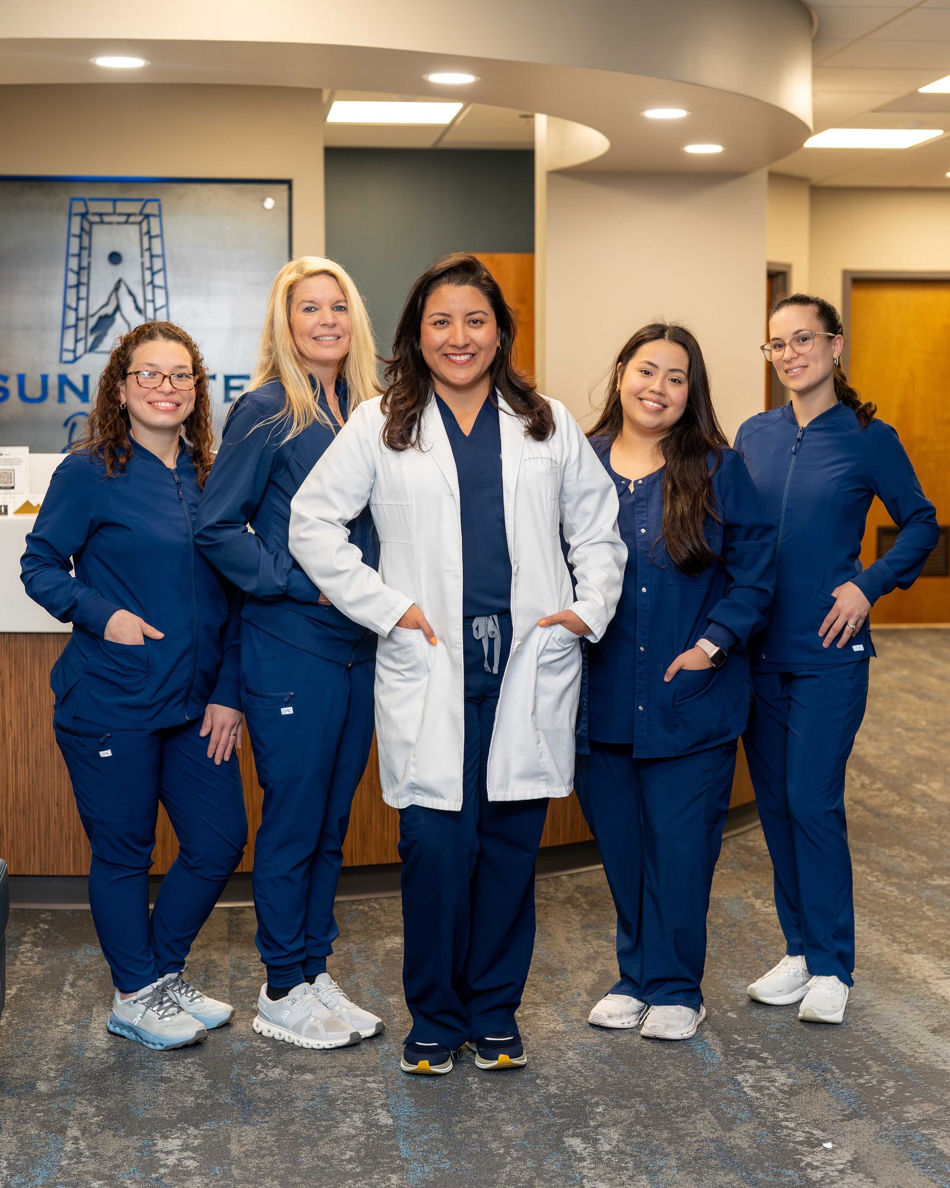 Five healthcare workers in blue scrubs and white coats pose in a hospital lobby.