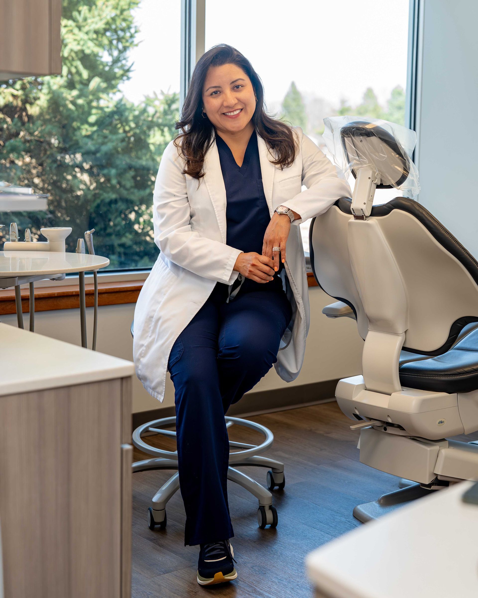 Dentist in white coat sitting on stool beside dental chair in a bright clinic room