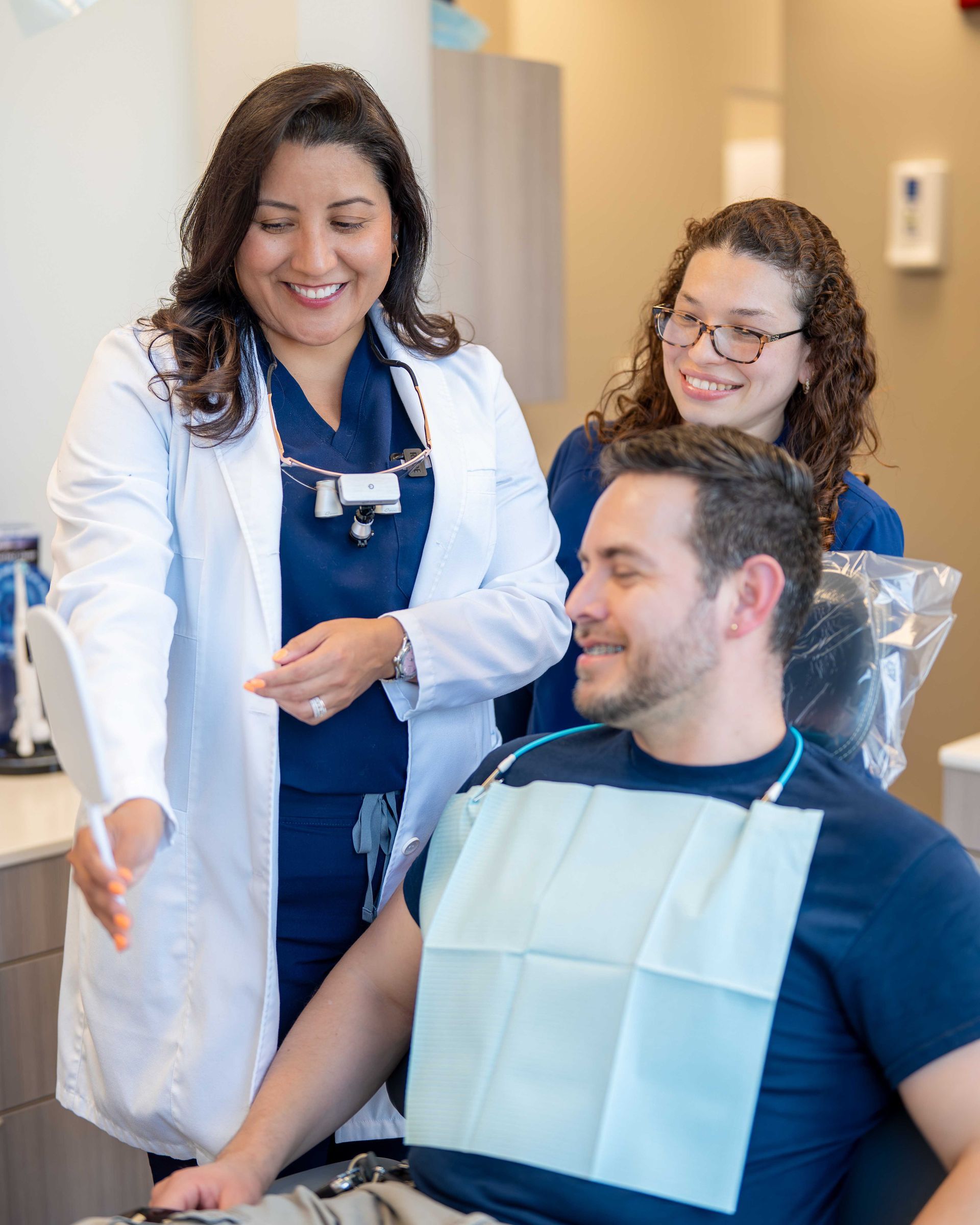 Dentist and assistant smiling with a seated patient in a dental clinic, wearing a bib and discussing treatment.