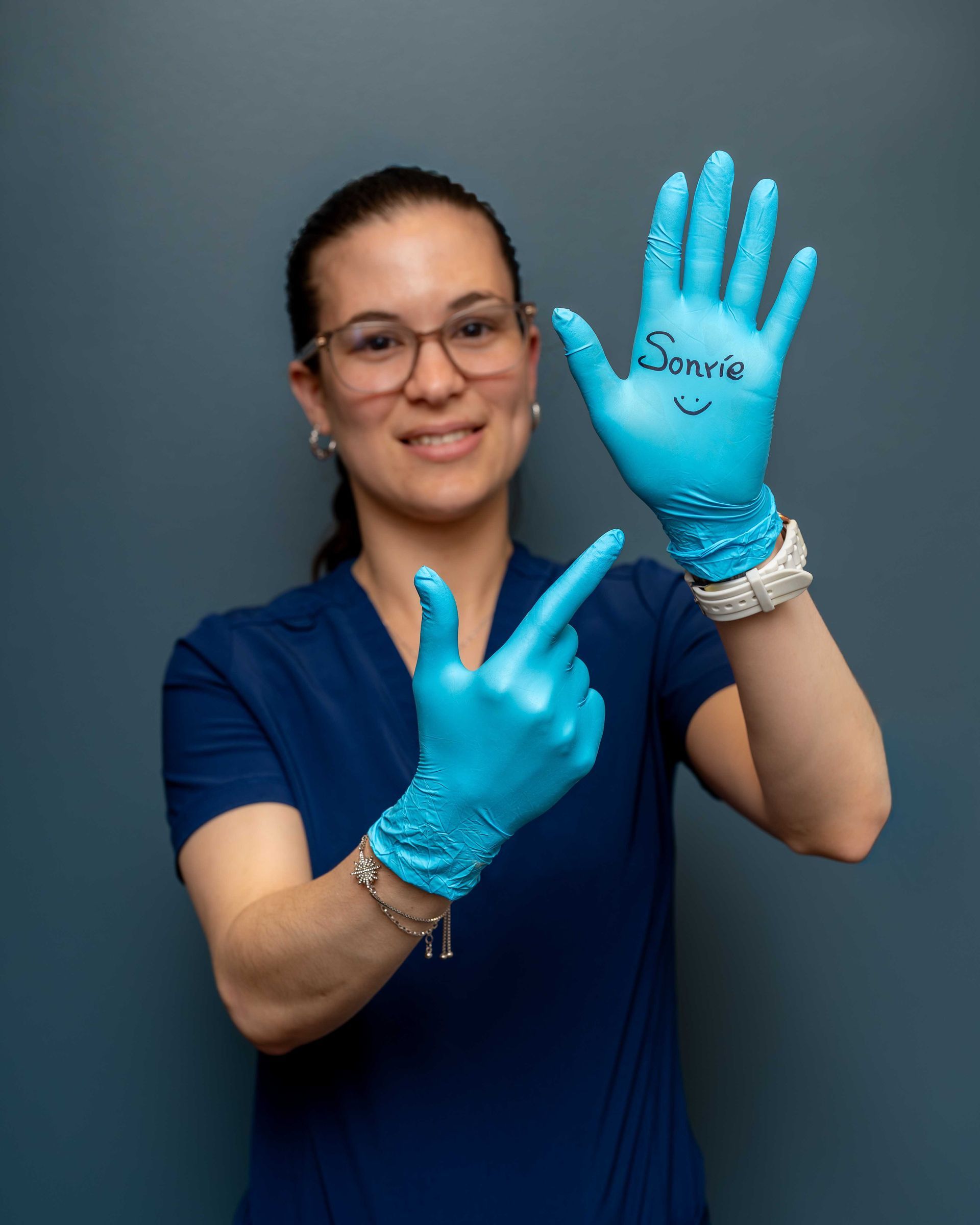 Smiling person in blue gloves and scrubs holds up hands to show a ring and painted nails.