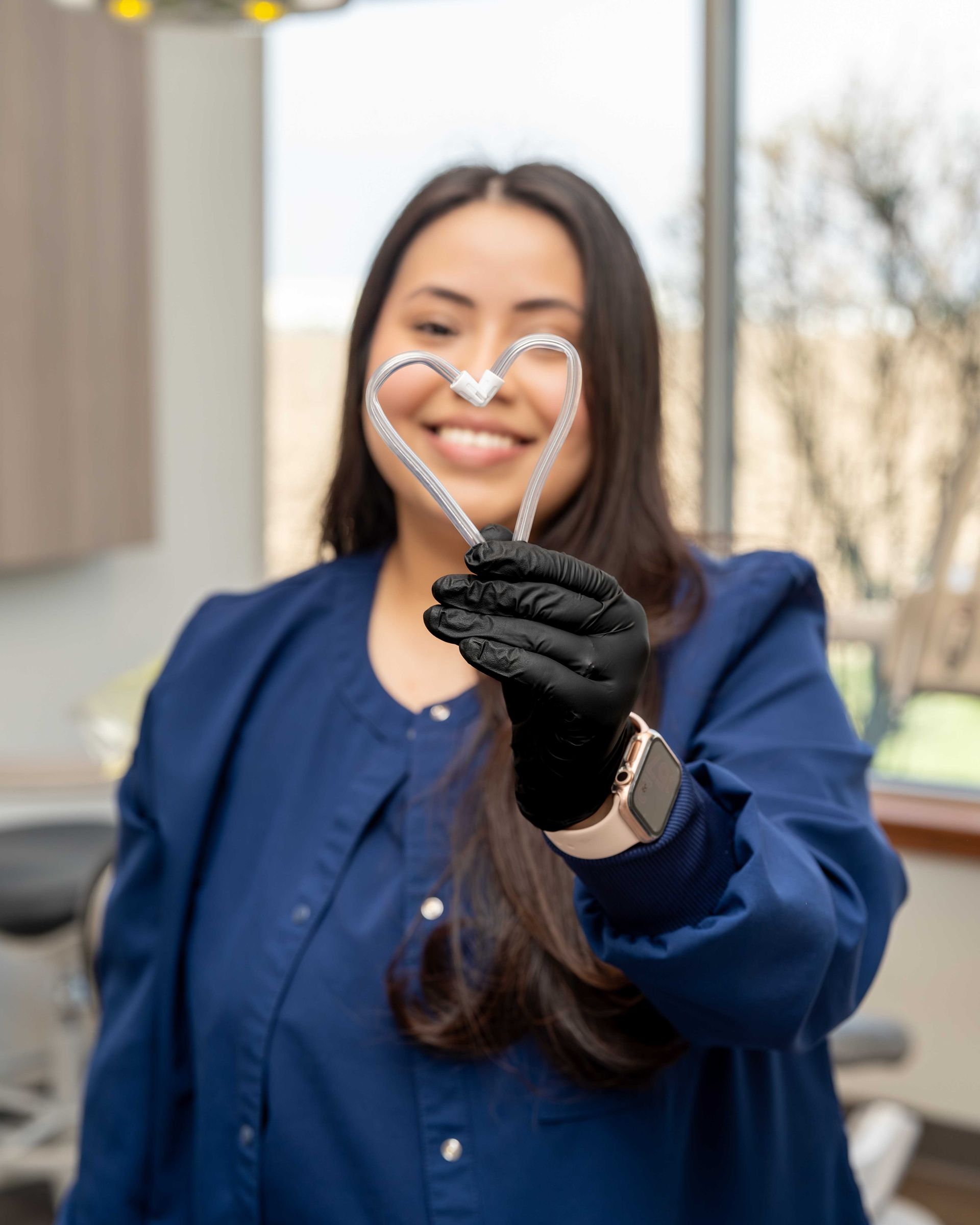Person in blue blouse and black gloves holds a heart-shaped object, smiling outdoors.