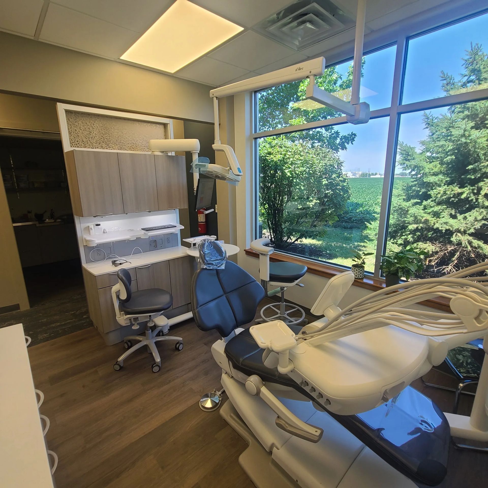 Dental exam room with chair, cabinets, and large windows overlooking trees.