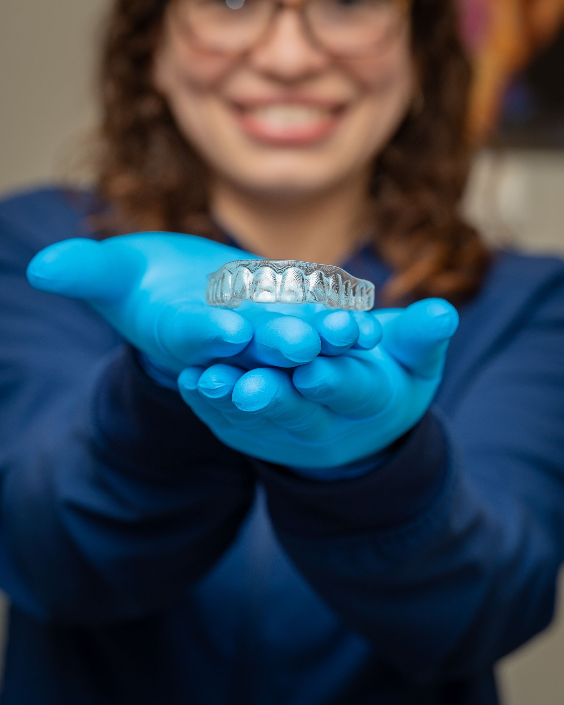 Gloved hands holding a clear dental aligner, with a smiling person blurred in the background