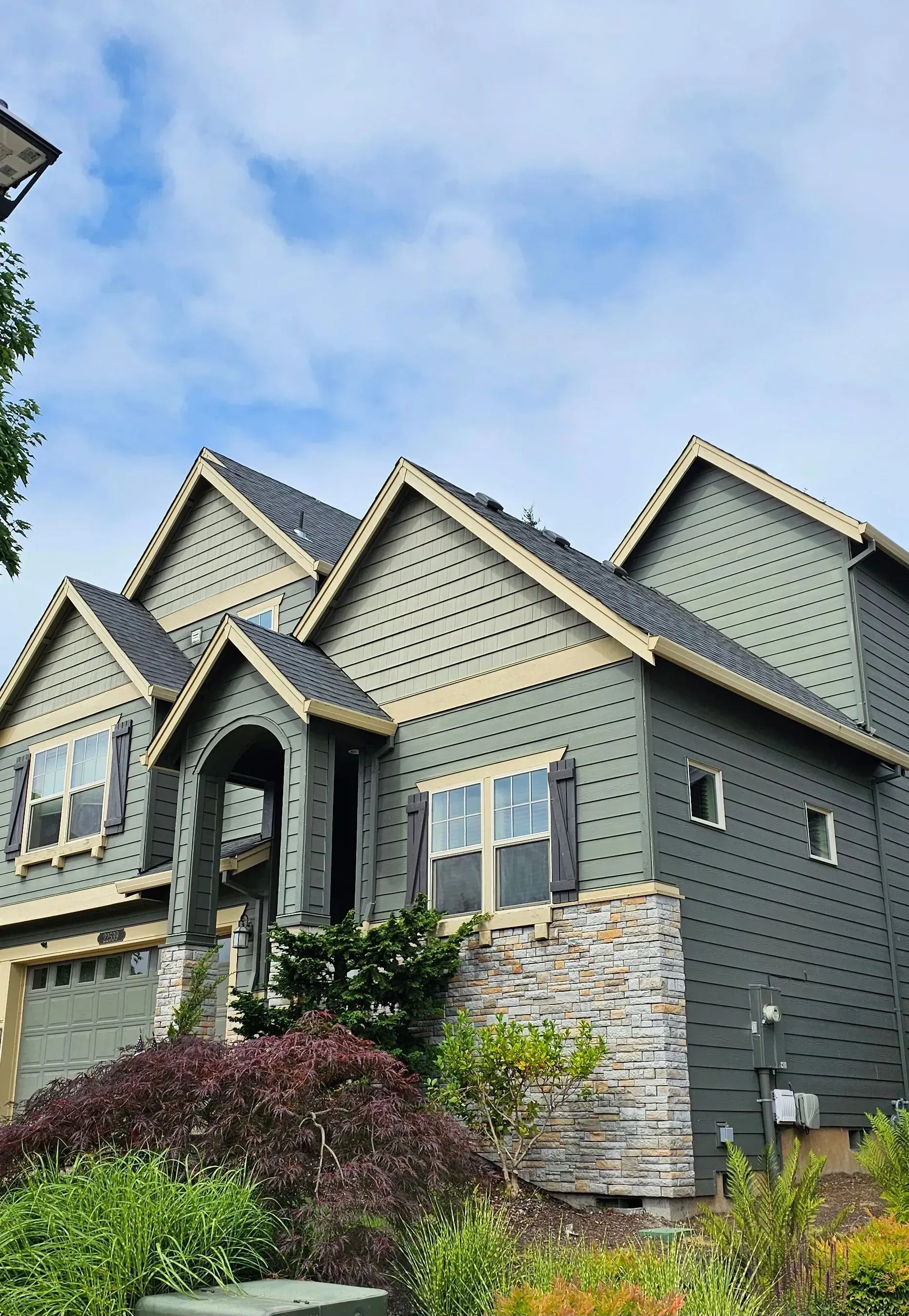 Two-story gray house with gabled roofs, stone accents, and landscaped shrubs under a blue sky