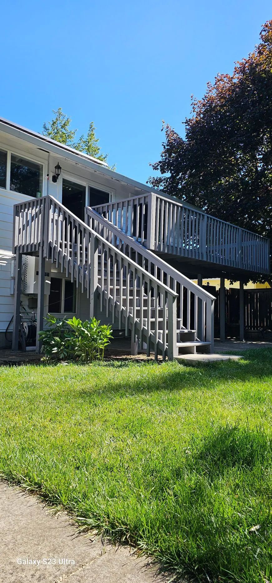Suburban house with white stairs and railing, green lawn, and trees under a bright blue sky