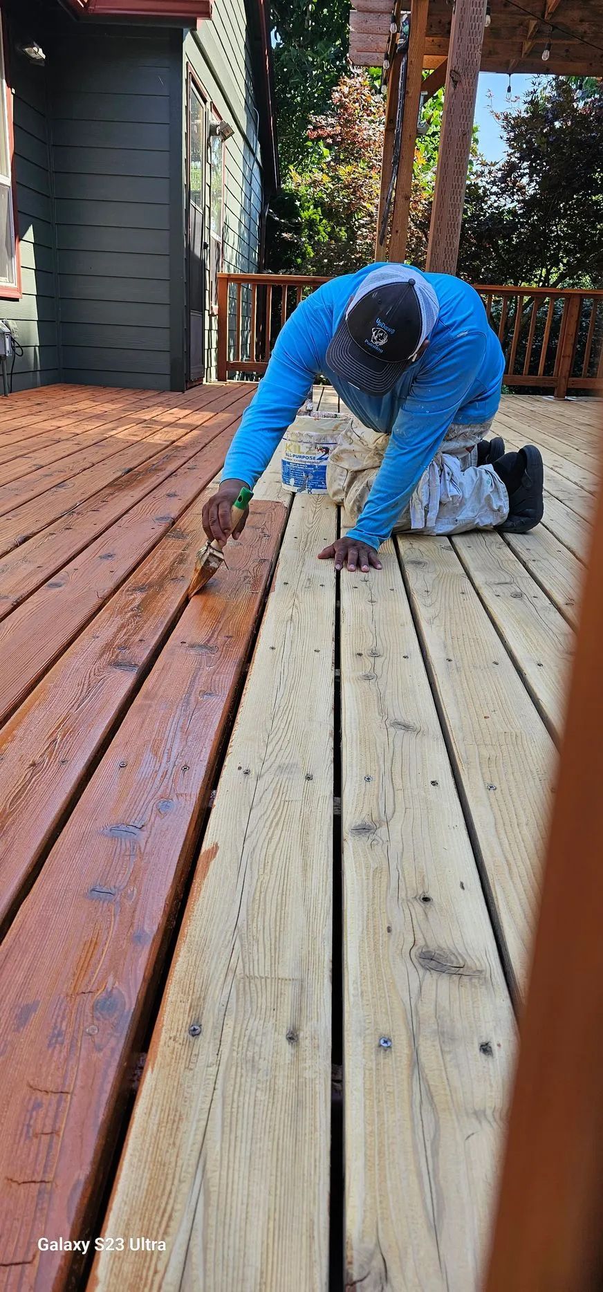 Person in a blue jacket kneels on a wooden deck, working on weathered floorboards with a tool.