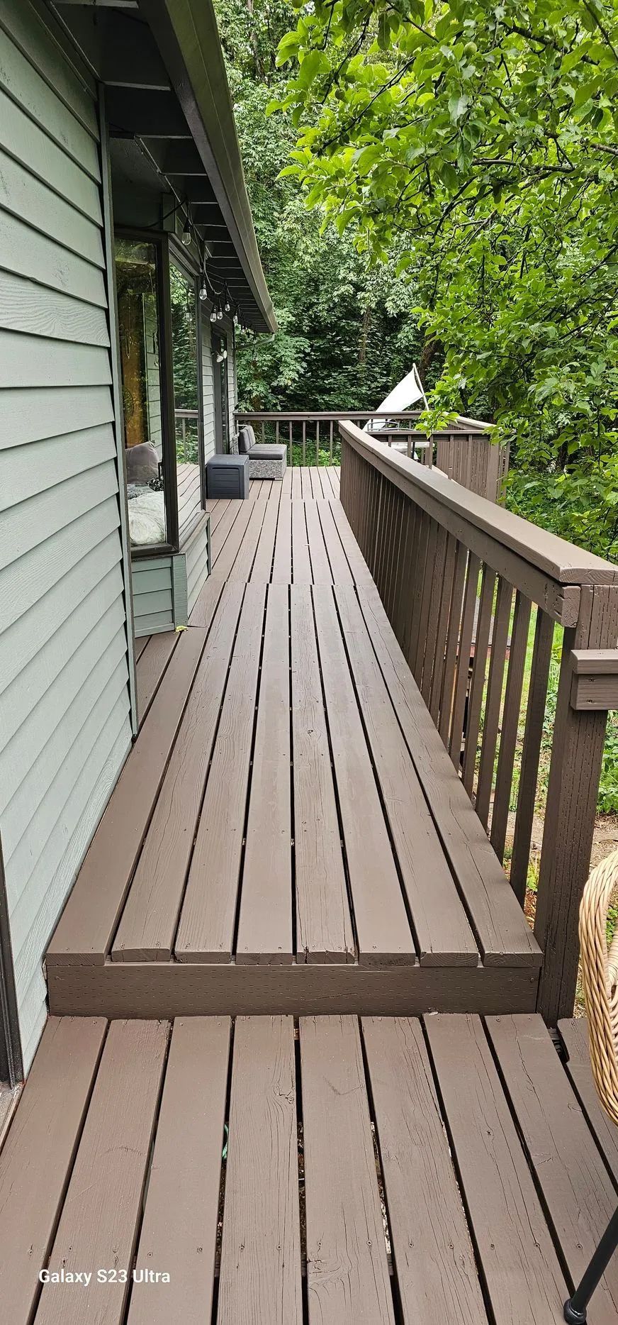 Wooden deck walkway beside a house, bordered by a railing and leafy green trees.