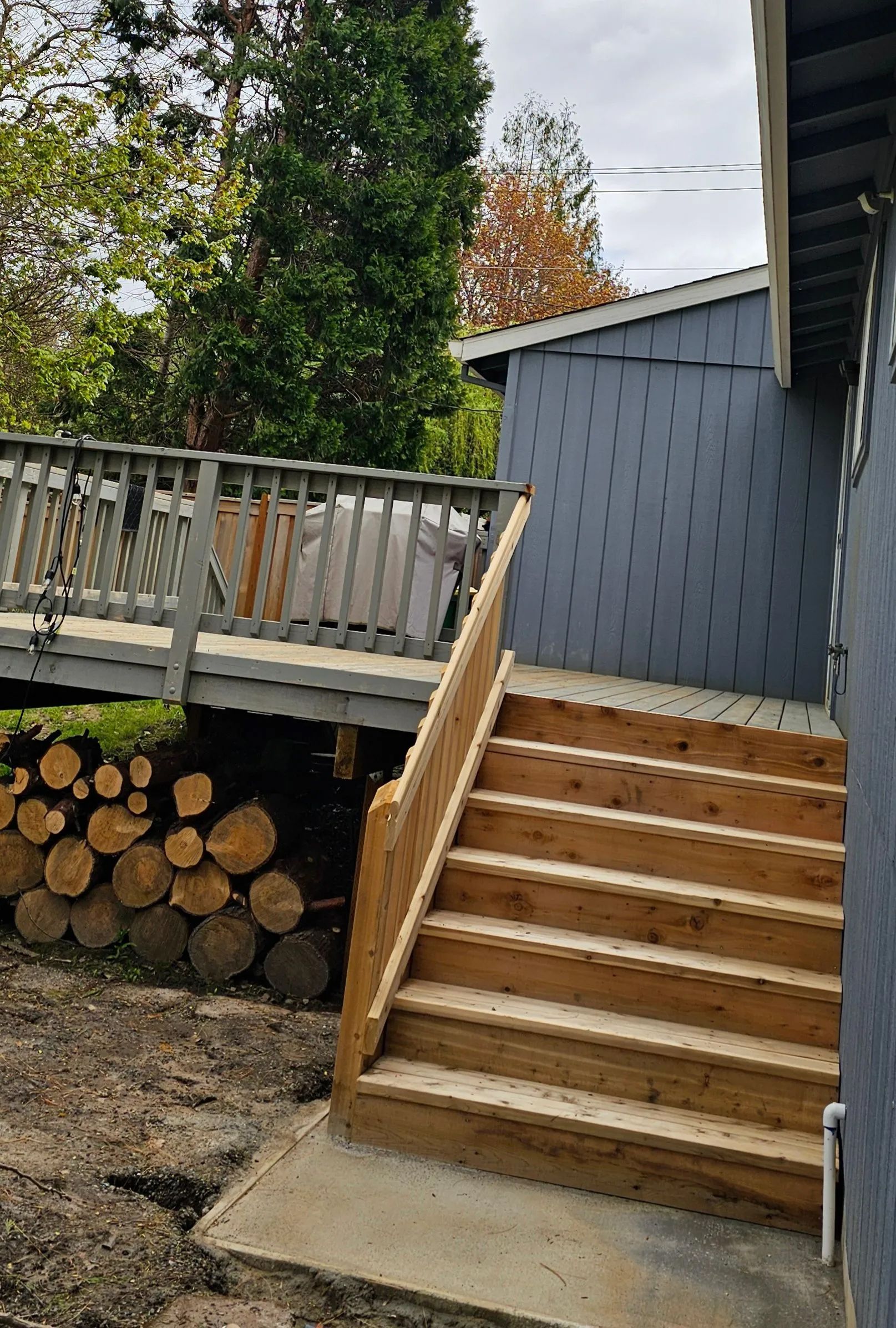 Outdoor wooden stairs leading to a deck beside a gray house, with stacked firewood underneath.