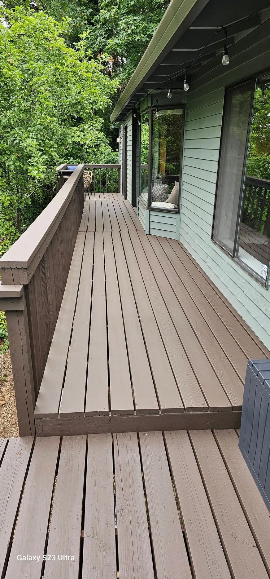 Narrow wooden deck walkway beside a green house, overlooking trees and a hillside