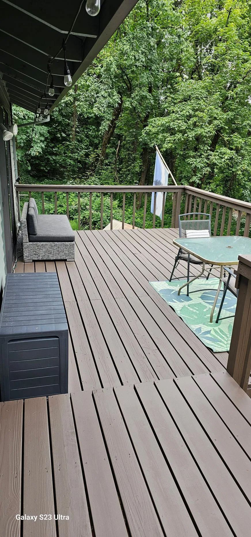 Wooden deck with outdoor sofa, glass table, umbrella, and dense green trees beyond the railing.