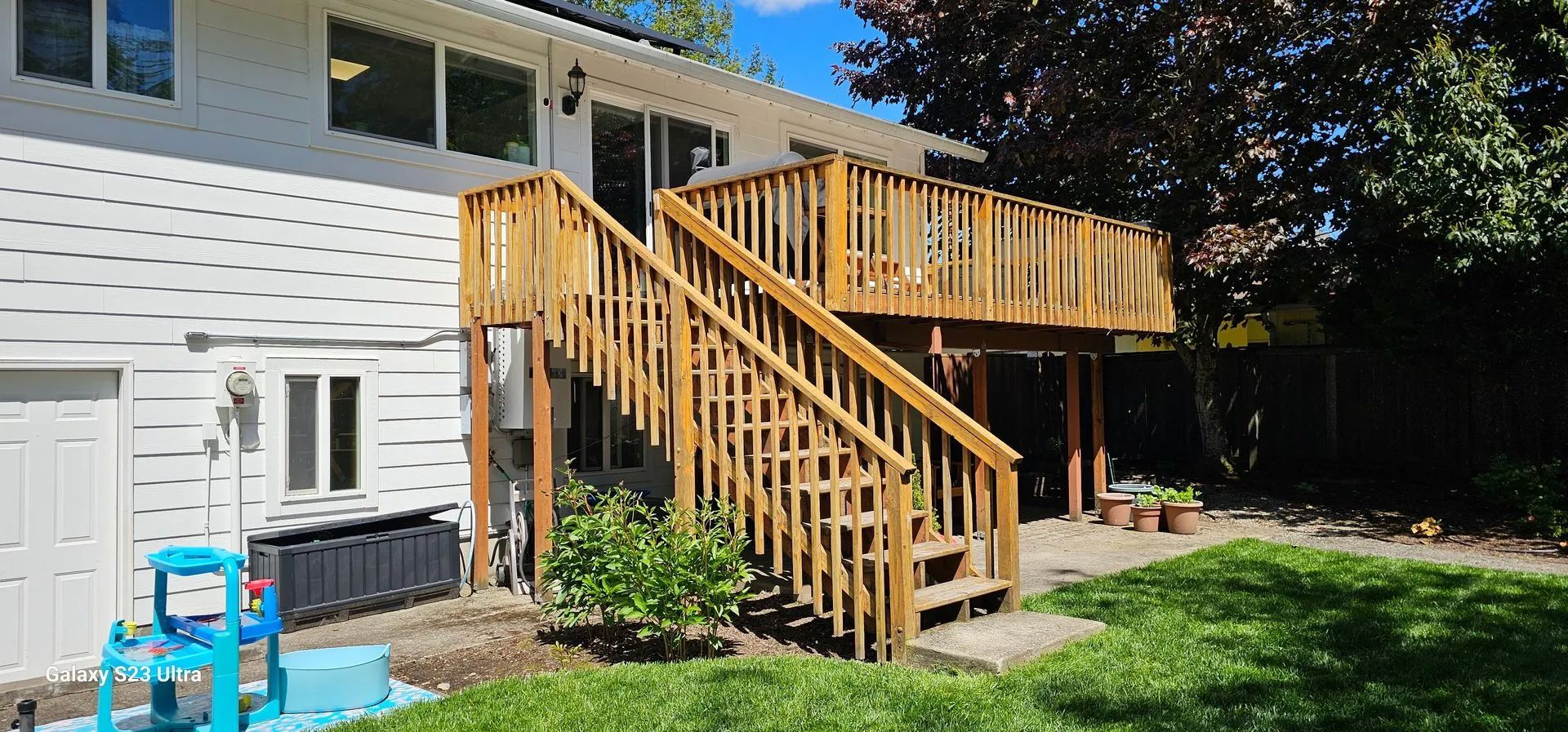 Wooden deck and stairs on the side of a white house in a sunny backyard.