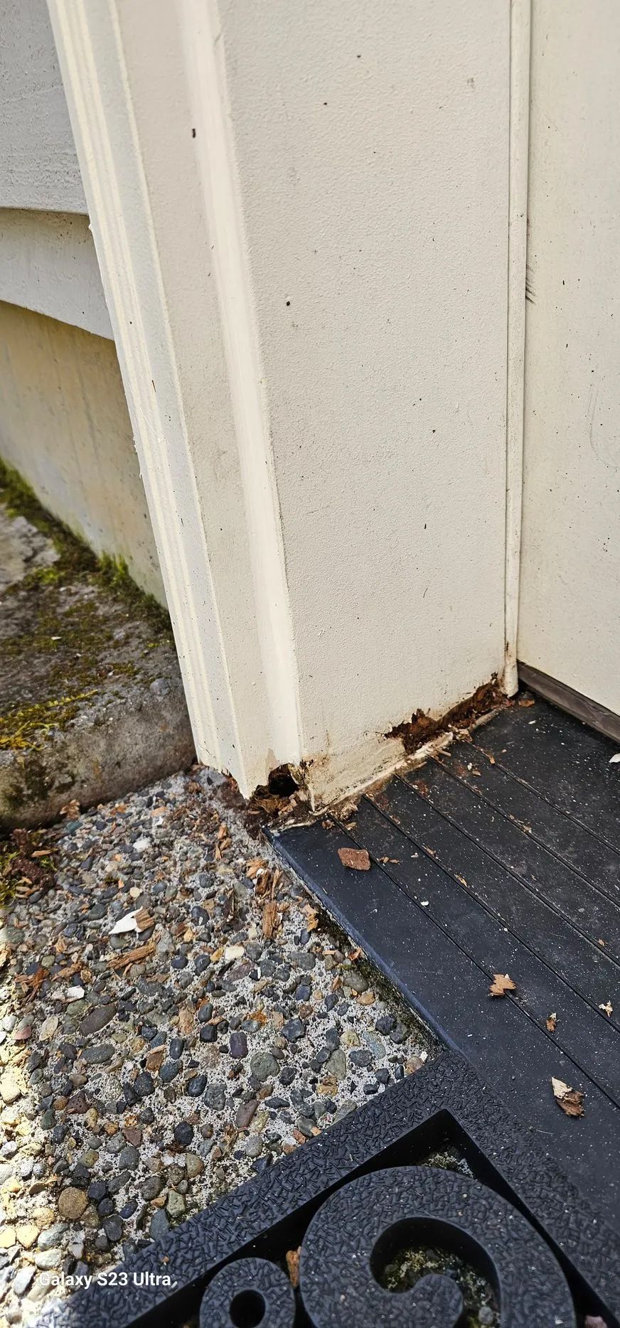 Close-up of a white doorframe and threshold with chipped base, gravel, and dark doormat tiles.