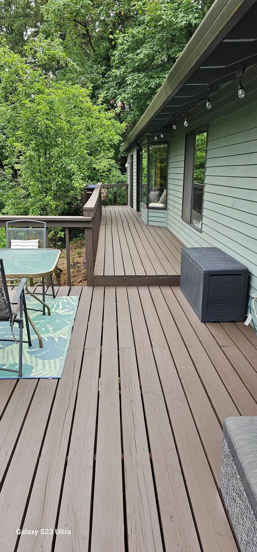 Wooden deck beside a house, with railing, string lights, patio furniture, and trees beyond.