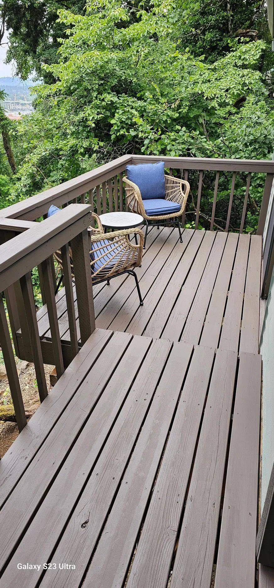 Wooden deck with a blue cushioned chair, small table, and lush green trees beyond the railing.