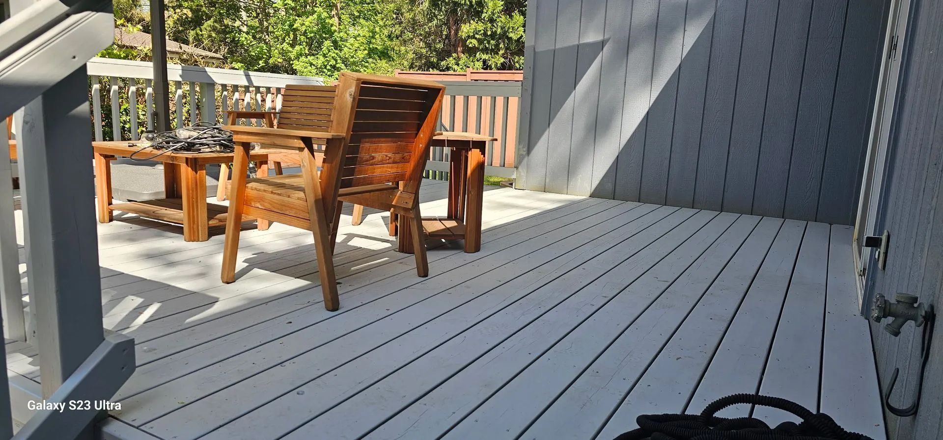 Wooden patio table and chairs on a gray deck in bright sunlight, with trees and a fence nearby