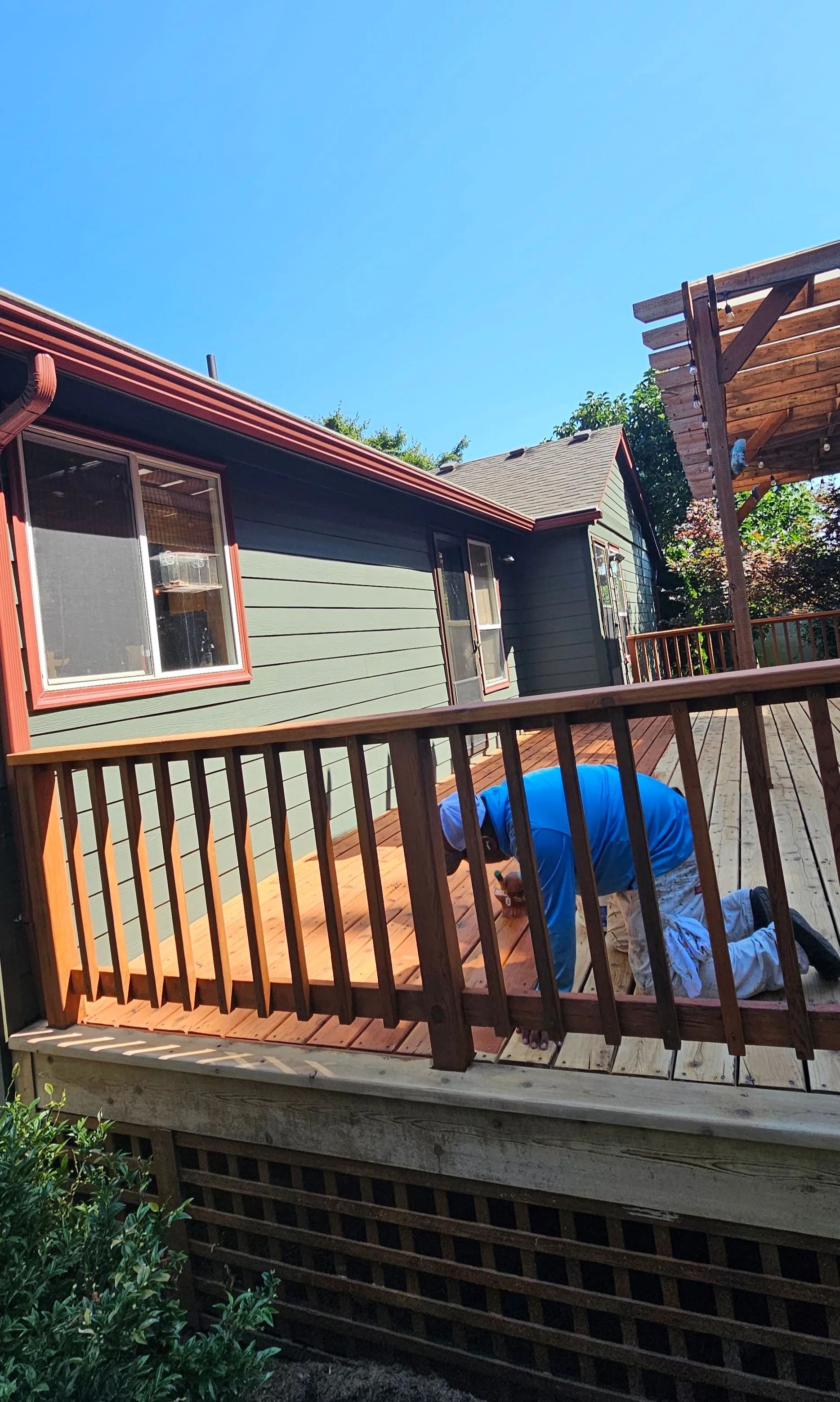 Person bending over on a wooden deck beside a gray house with a red roof and blue sky