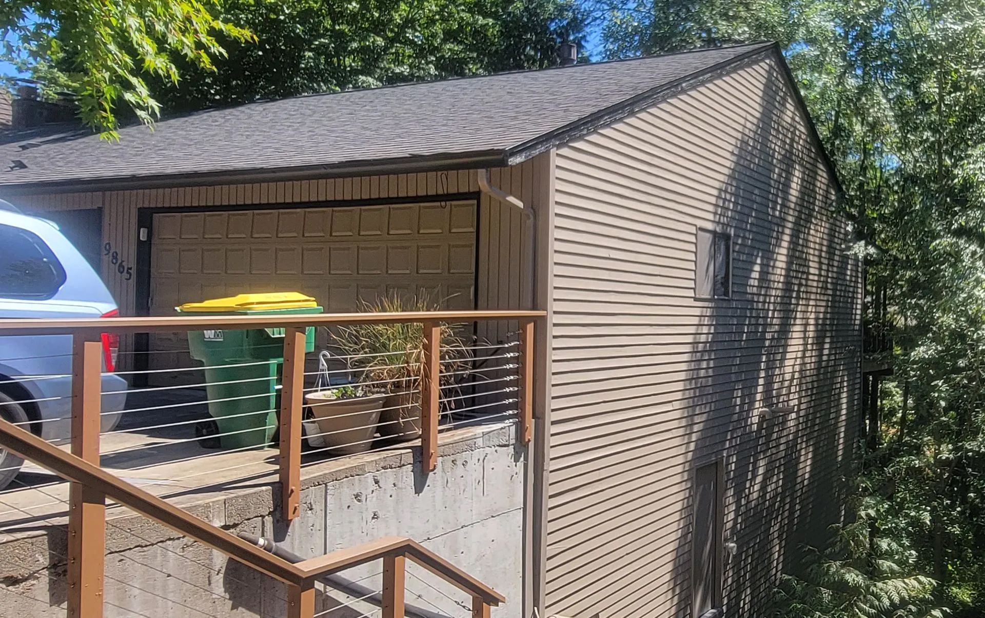 Wooden house exterior with a sloped roof, railing, and a green trash bin beside the wall
