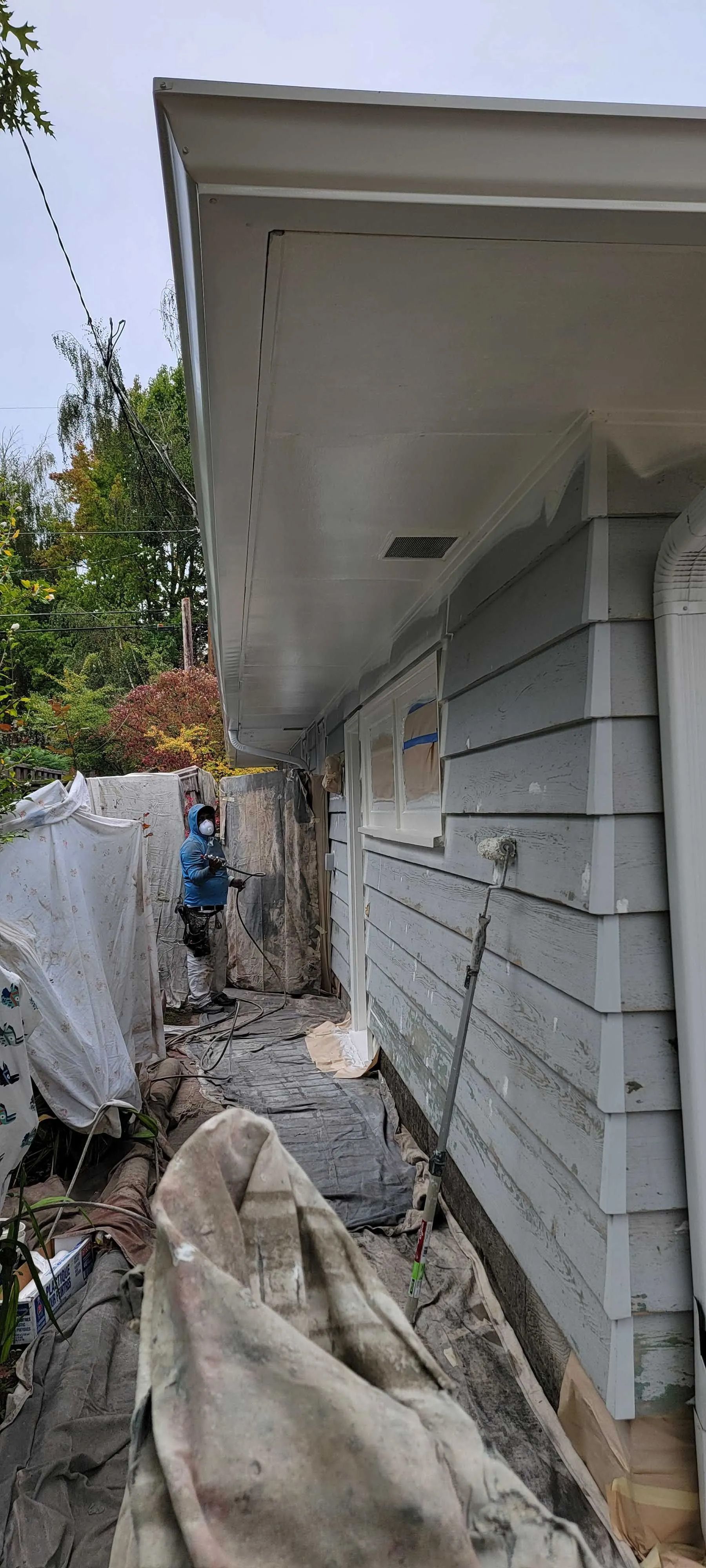 Narrow side yard beside a white house with construction debris and covered materials on the ground.