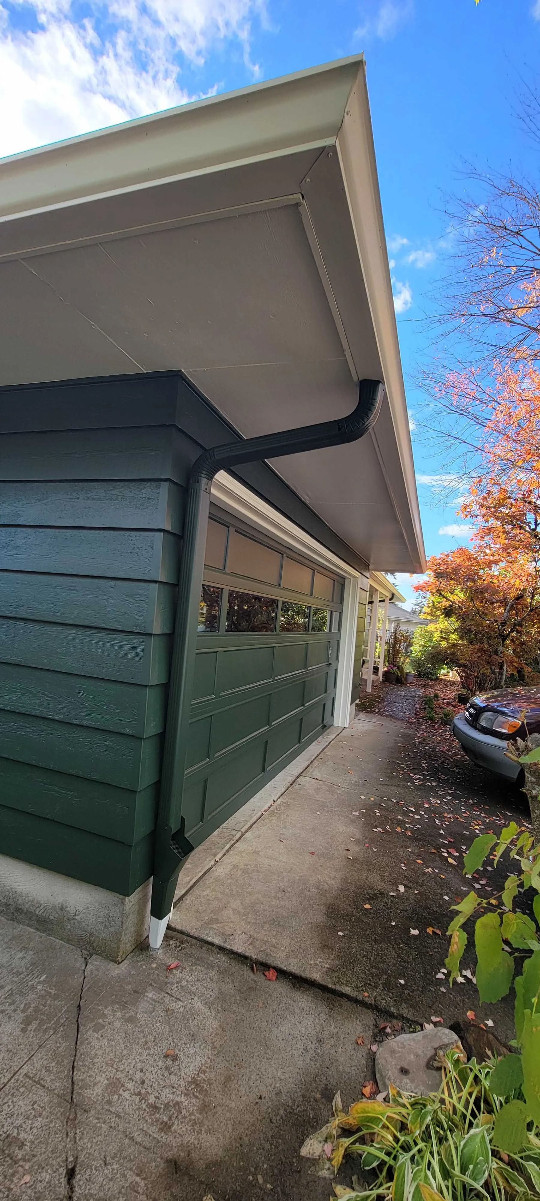 Side view of a green house exterior with a concrete walkway, garage eaves, and parked car beside plants.