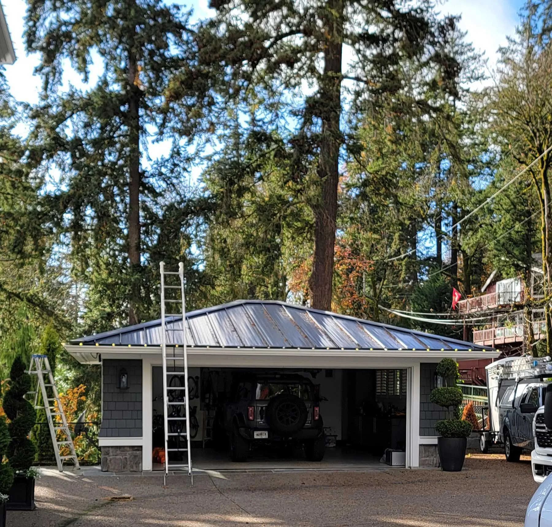 Detached garage with open bays and a car, surrounded by tall trees in a wooded driveway