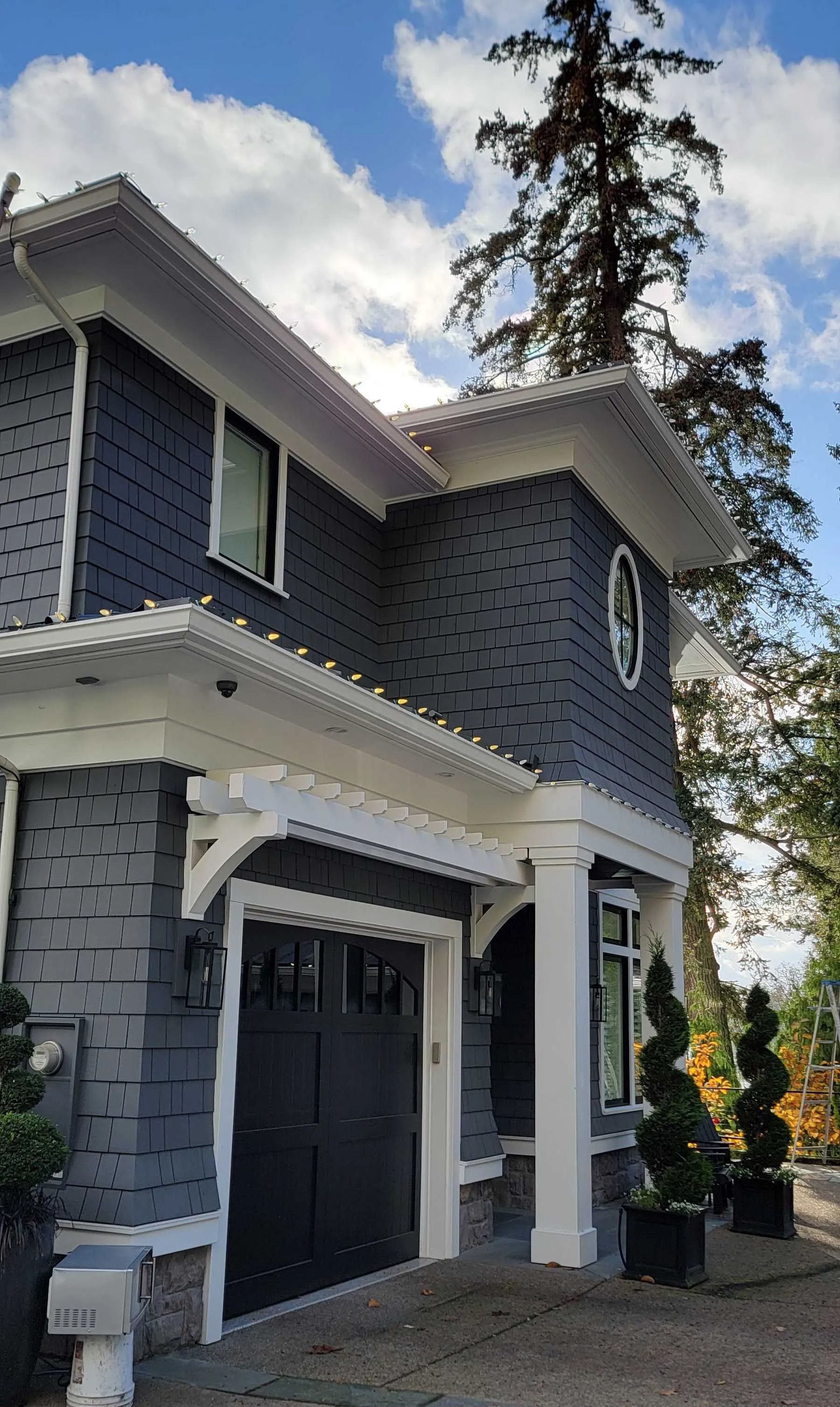 Gray two-story house with white trim, black garage door, and tall evergreen trees in the yard