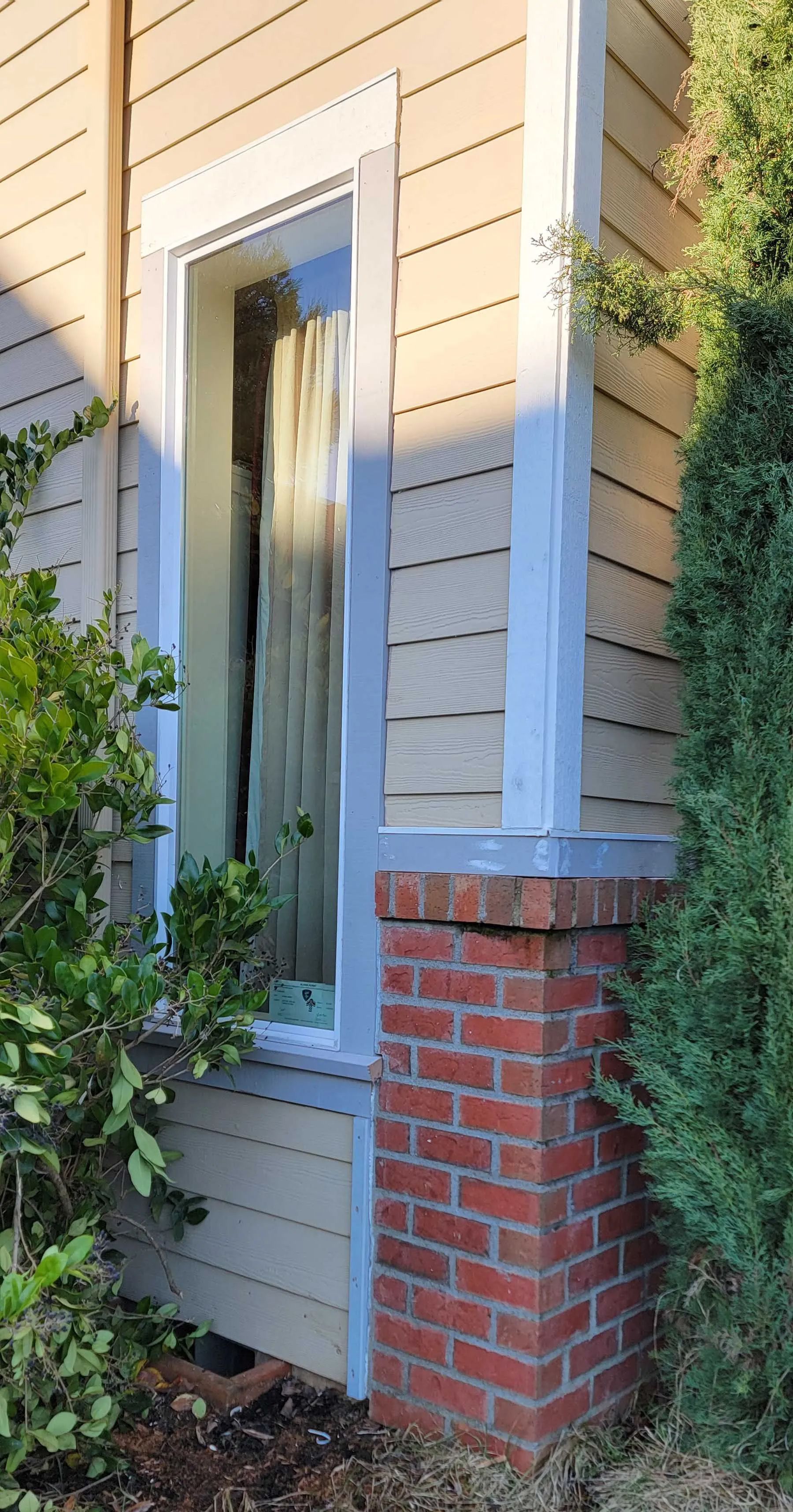 Front corner of a beige house with white trim, brick column, and a narrow window partly covered by plants