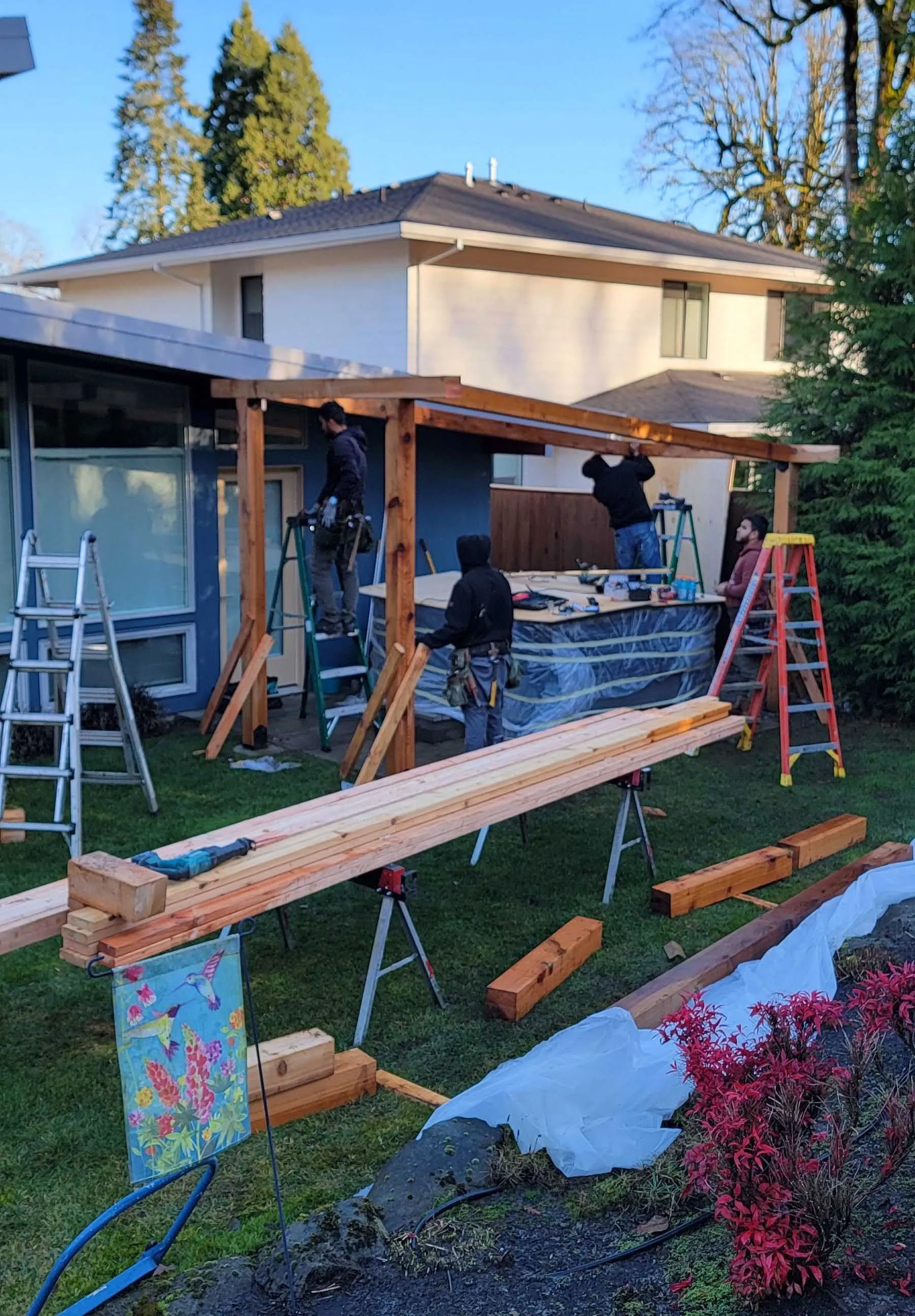 Home exterior under construction with wooden beams, ladders, and workers building a covered patio