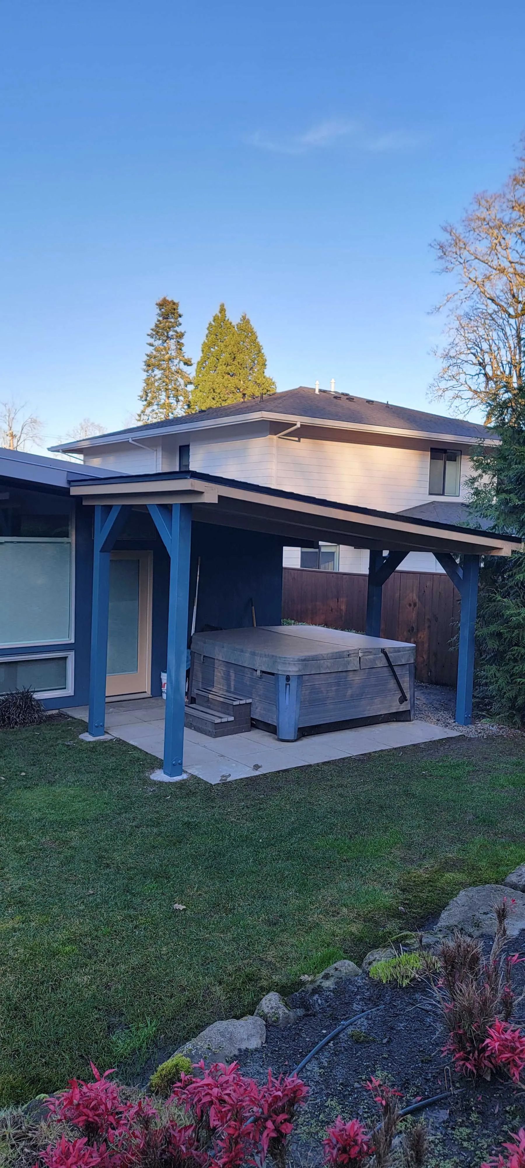 Backyard gazebo with white columns and blue trim beside a lawn and flower bed at sunset