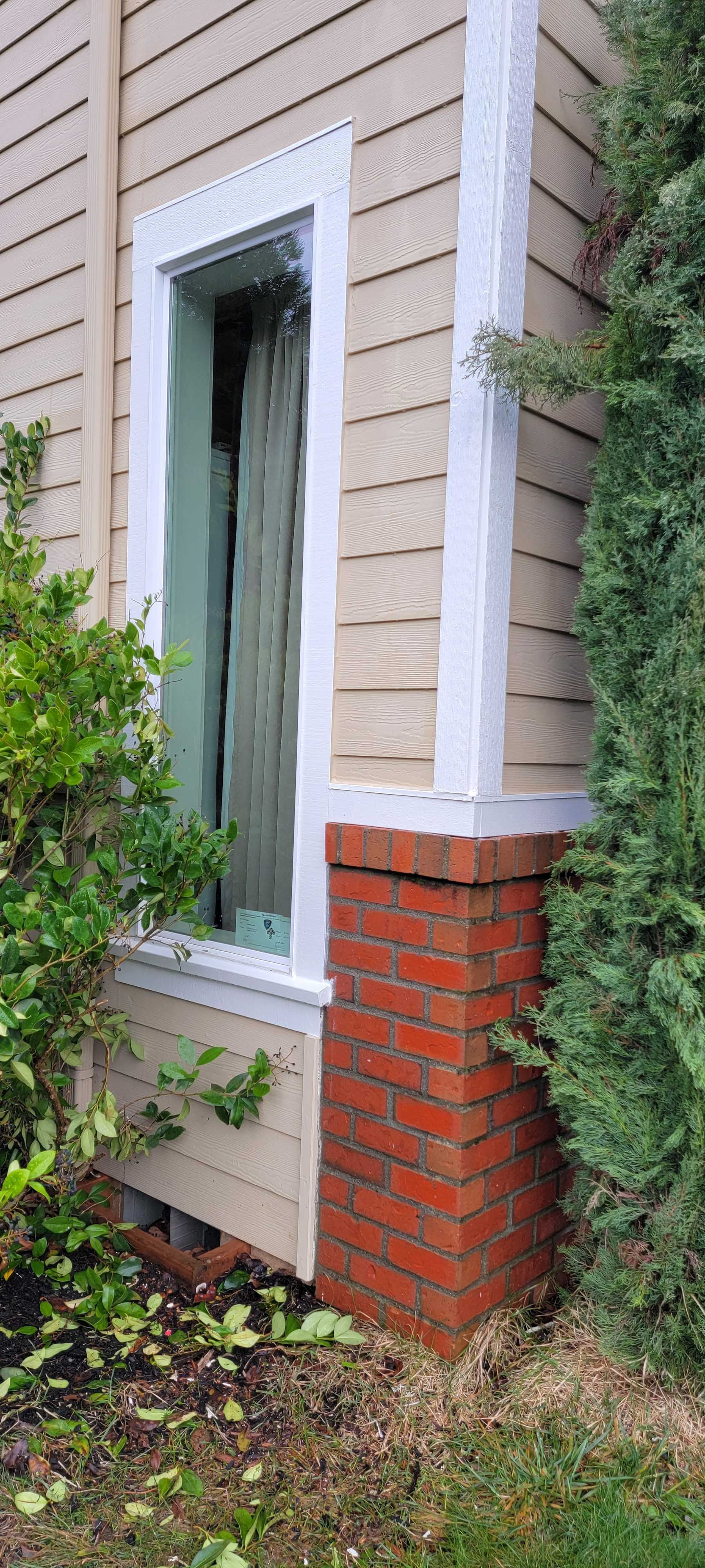 Narrow vertical window on a beige house with white trim and a red brick base, framed by greenery.