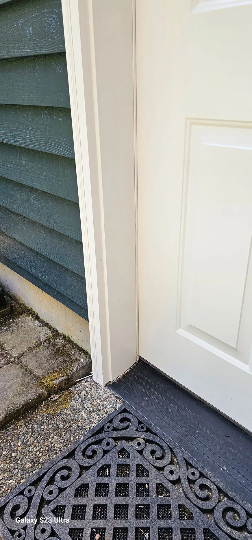 Front door corner with white trim, blue siding, and a black lattice doormat on stone pavers