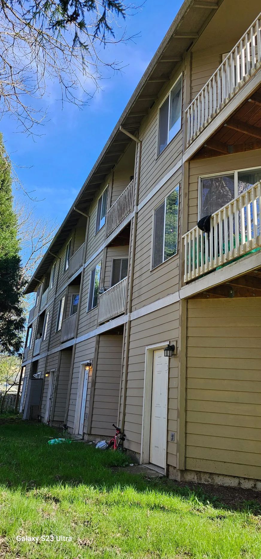 Three-story beige apartment building with balconies and white railings beside a grassy lawn.