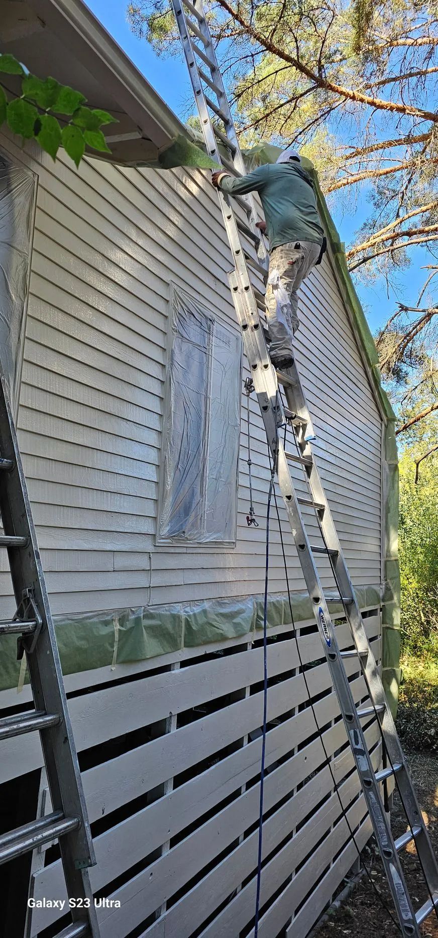 Person on a ladder painting the exterior of a beige house beside a white fence