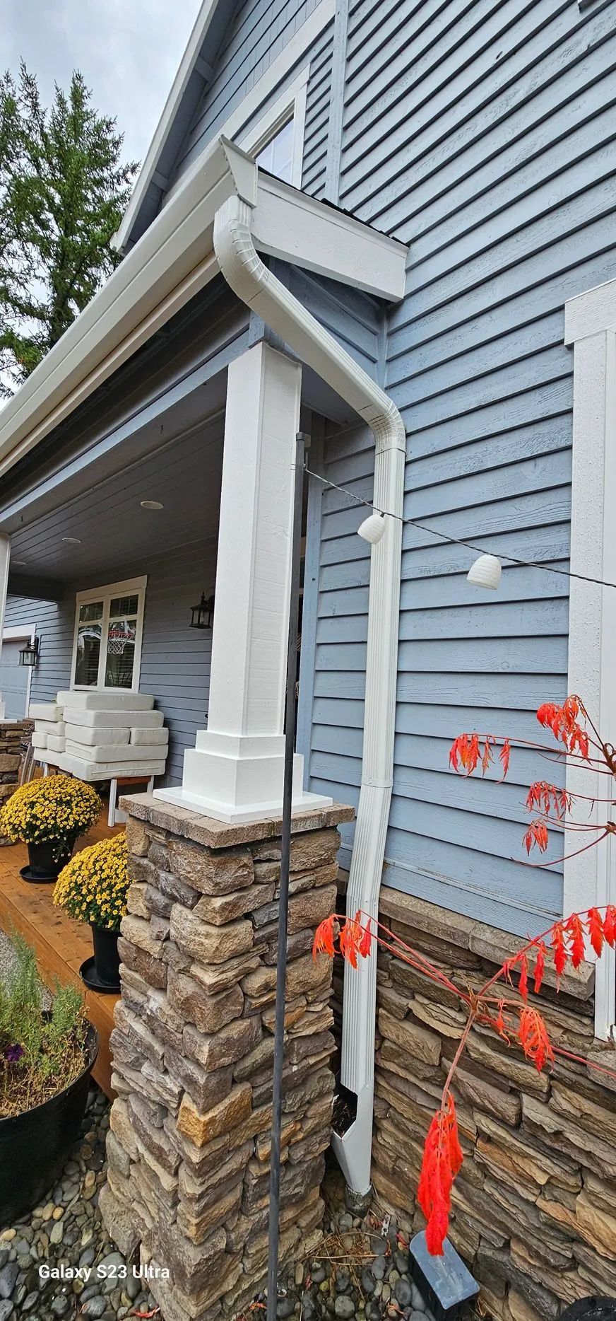 White downspout on blue house with stone foundation and white trim by the front porch
