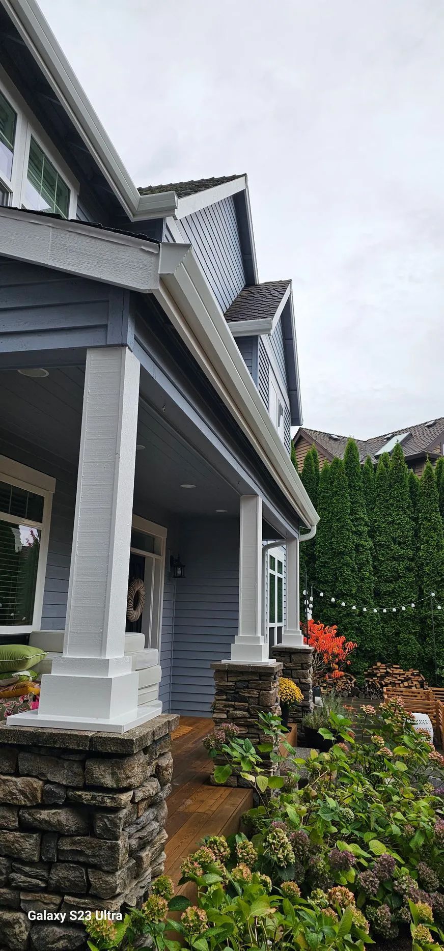 Gray house porch with white trim, stone pillars, and lush green shrubs under an overcast sky